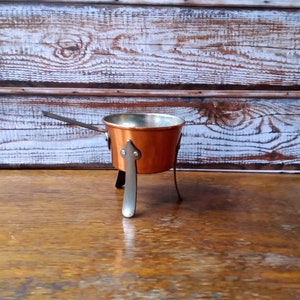Op de afbeelding: Een kleine koperen pot met een handvat en drie poten. De pot staat op een houten oppervlak.