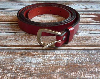 May include: A rolled-up red leather belt with a gold-colored buckle. The belt is on a wooden surface with a rustic, weathered appearance. The belt is a classic accessory.