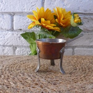 Puede incluir: Una pequeña olla de cobre con asa se asienta sobre un soporte de metal con tres patas. La olla está sobre una superficie de mimbre marrón. Un ramo de girasoles amarillos está en el fondo.