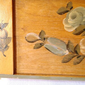 May include: A wooden tray with a painted floral design. The tray has a light brown wood background with a green border. The flowers are painted in white, light green, and brown.