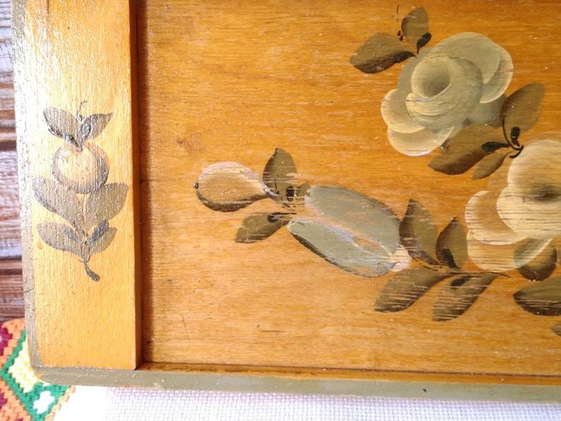 May include: A wooden tray with a painted floral design. The tray has a light brown wood background with a green border. The flowers are painted in white, light green, and brown.