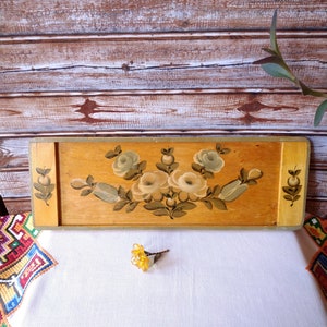 May include: A wooden tray with a painted floral design featuring white flowers and green leaves. The tray has a light brown wood finish and a gold trim.