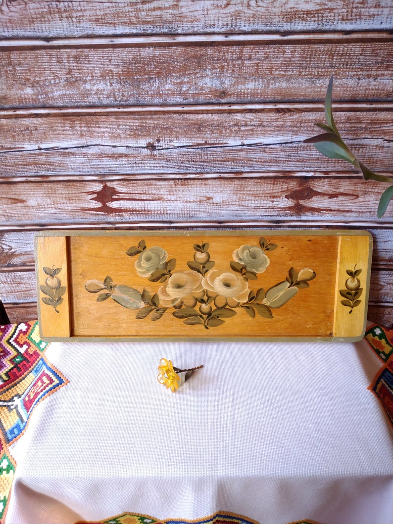 May include: A wooden tray with a painted floral design featuring white flowers and green leaves. The tray has a light brown wood finish and a gold trim.