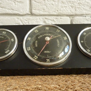 May include: A black wooden board with three round analog gauges. The gauges are labeled "Hygro", "Thermo", and "For Anderlug" with the text "Regn" and "Smukt" on the gauge labeled "For Anderlug".
