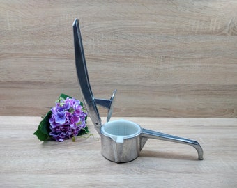 May include: A silver potato ricer with a white plastic bowl and a lever handle. The ricer is on a wooden surface with a purple flower bouquet.