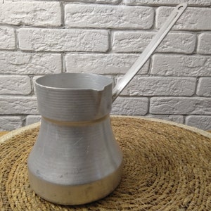 Peut inclure: Une cafetière en aluminium argentée avec une longue poignée. La cafetière est posée sur un tapis en paille tressée.