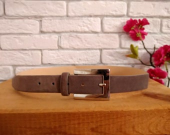 May include: A gray suede belt with a silver buckle. The belt is on a wooden surface.
