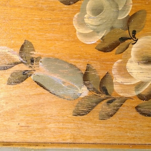 May include: A wooden surface with a painted floral design. The flowers are white with green leaves and are painted in a rustic style.