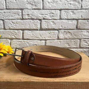 May include: A brown leather belt with a silver buckle. The belt has a stitched design and text along the bottom. The belt is displayed on a wooden surface with a brick background.