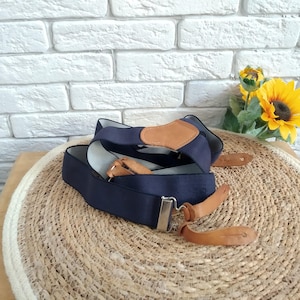 Puede incluir: Un par de tirantes azul oscuro con correas de cuero marrón y una hebilla plateada. Los tirantes están sobre una estera de paja tejida.