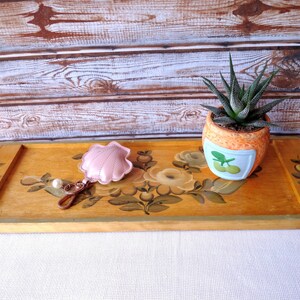May include: A wooden serving tray with hand-painted floral designs in shades of yellow, green, and white. A small pink seashell keychain and a potted succulent plant are on the tray.