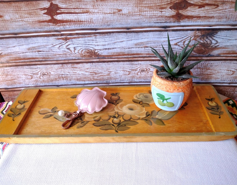 May include: A wooden serving tray with hand-painted floral designs in shades of yellow, green, and white. A small pink seashell keychain and a potted succulent plant are on the tray.