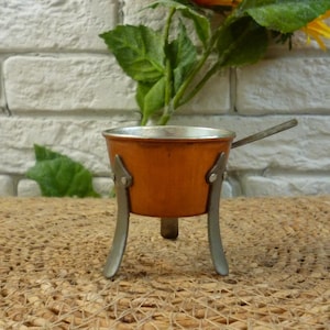 May include: A small, copper-colored pot with a wooden handle sits on a woven mat. The pot has three legs and is made of metal.