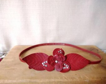 May include: A red headband with a floral and leaf design. The headband features a central flower with rhinestone accents and two textured leaves on either side. The band is thin and flexible.