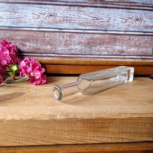 Può includere: Una bottiglia di vetro trasparente con forma quadrata e collo arrotondato. La bottiglia è appoggiata su una superficie di legno.