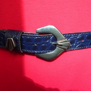 以下が含まれることがあります： シルバーのバックルとテクスチャード加工の模様が付いたネイビーブルーのベルト。赤い衣服の上から着用されています。バックルはユニークな形状とデザインをしています。