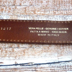 Puede incluir: Primer plano del interior de un cinturón de cuero marrón, mostrando texto en relieve. El texto incluye "ART. 111217", "VERA PELLE - GENUINE LEATHER", "FATTA A MANO - HAND MADE", "MADE IN ITALY" y la talla "95/110".