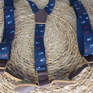 Puede incluir: Tirantes azules con un patrón de calavera y huesos cruzados blancos y detalles rojos. Los tirantes tienen correas ajustables de cuero marrón y clips.