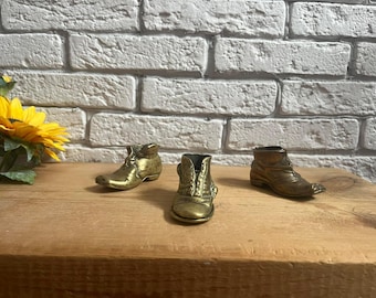 Puede incluir: Tres pequeñas botas de latón de estilo antiguo están dispuestas sobre una superficie de madera. Las botas están detalladas con cordones y costuras, y tienen un aspecto vintage y desgastado. Un girasol amarillo es visible en el fondo contra una pared de ladrillos blancos.