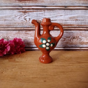 Peut inclure: Un petit pichet en céramique brun avec un motif de fleur blanche. Le pichet a un bec verseur et une anse. Il est posé sur une surface en bois.