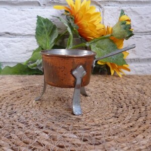 Può includere: Una piccola pentola di rame con un manico e tre gambe. La pentola è appoggiata su un tappetino intrecciato.