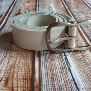 Peut inclure: Une ceinture en cuir beige clair enroulée, avec une boucle carrée de couleur argentée. La ceinture est posée sur une surface en bois avec un motif de grain de bois naturel. La boucle est ouverte.