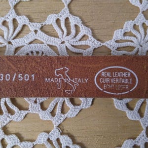 Puede incluir: Un cinturón de cuero marrón con el texto "30/501" y "Made in Italy". El cinturón mide 76 cm de largo y está etiquetado como "Real Leather". El fondo es un tapete de encaje blanco.
