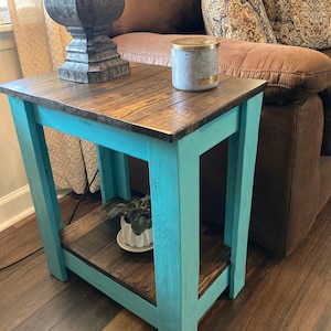 May include: A teal blue end table with a wooden top and a lower shelf. The table has a distressed wood finish and is decorated with a candle and a potted plant.