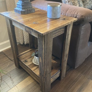 May include: A rustic wooden end table with a lower shelf. The table is stained a dark brown color and has a distressed finish. It is sitting next to a brown leather couch with a decorative pillow.