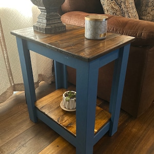 May include: A small wooden end table with a blue painted frame and a natural wood top. The table has a lower shelf and is decorated with a small potted plant.