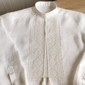 May include: A white linen shirt with embroidered details and a tassel. The embroidery is a geometric pattern in a lighter shade of white.