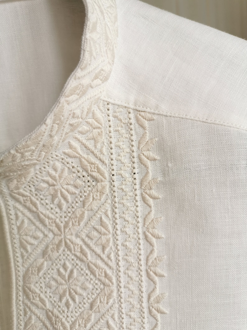 May include: White linen shirt with intricate embroidered details around the collar and neckline.
