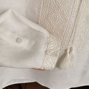 May include: A white linen shirt with embroidered details. The shirt has a button closure and a tassel on the sleeve.