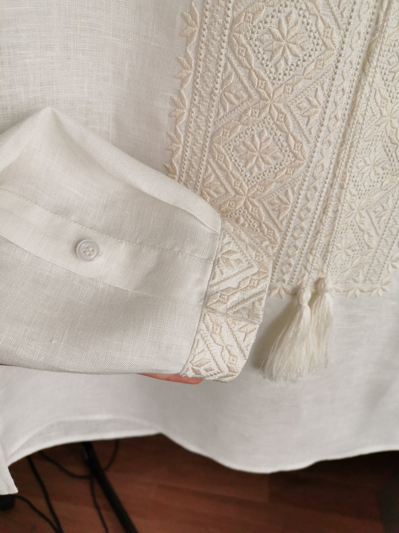 May include: A white linen shirt with embroidered details. The shirt has a button closure and a tassel on the sleeve.