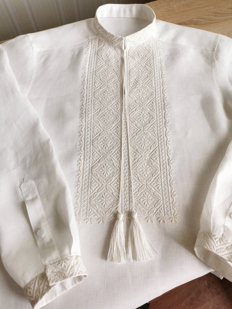 May include: A white linen shirt with embroidered details and a tassel. The embroidery is a geometric pattern in a lighter shade of white.