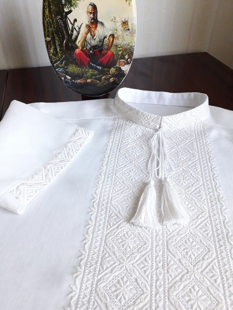 May include: White embroidered shirt with a traditional Ukrainian design. The shirt has a stand-up collar and long sleeves. The embroidery is done in white thread and features a geometric pattern.
