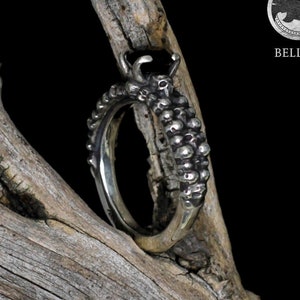 Könnte beinhalten: Ein silberner Ring mit einem schwarzen Edelstein und einem strukturierten, organischen Design. Der Ring wird auf einem Stück Treibholz präsentiert.