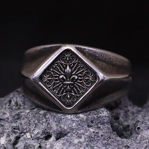 May include: A silver ring featuring a fleur-de-lis emblem within a diamond-shaped setting. The ring has a broad band and a slightly weathered appearance. The ring is displayed on a grey, textured surface, suggesting a vintage aesthetic.