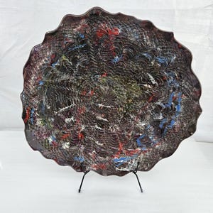 May include: A large, round ceramic platter with an abstract design. The platter has a wavy edge and is decorated with a swirling pattern of black, red, blue, and green paint.
