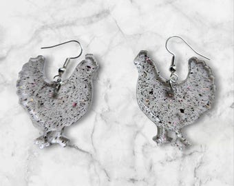 May include: A pair of silver-colored chicken-shaped earrings. The earrings are made of a speckled material with silver-colored ear wires. The chicken earrings have a glittery appearance and are set against a white marble background.