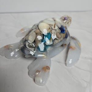 May include: A decorative sea turtle figurine crafted from clear, glitter-infused resin. The shell is filled with an assortment of small, natural seashells in shades of white, tan, and blue, creating a textured, multi-dimensional appearance.