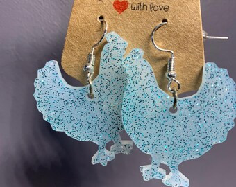 May include: A pair of light blue chicken-shaped earrings featuring a glittery finish and silver-toned metal hooks. The earrings are attached to a brown cardboard display card that has the text Handmade with love printed on it.