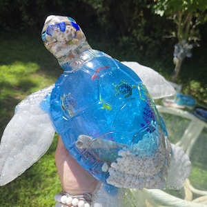 May include: A decorative sea turtle figurine crafted from clear and blue resin. The head is filled with small seashells, while the shell contains miniature turtle figures, colorful coral accents, and white pebbles embedded within the translucent blue material.