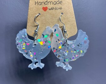 May include: A pair of chicken-shaped earrings made of translucent white resin with embedded iridescent holographic flakes. The earrings hang from silver-toned metal hooks and are displayed on a brown cardboard tag labeled Handmade with love.