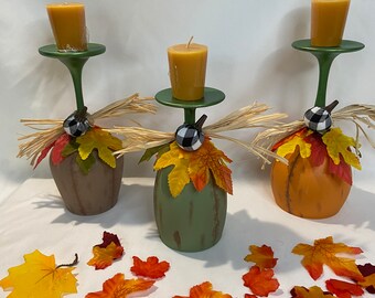 May include: Three decorative candle holders shaped like pumpkins, in shades of brown, green, and orange. Each holder has a green stem and a small candle. The pumpkins are adorned with fall leaves and a black and white checkered accent.