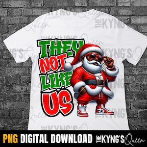 Op de afbeelding: Een wit T-shirt met een grafisch ontwerp van een zwarte kerstman met een zonnebril en een gouden ketting. De tekst "They Not Like Us" is geschreven in groene en rode letters.