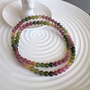 Pode incluir: Um colar de contas de pedras preciosas multicoloridas com contas verdes, rosa e amarelas. O colar é feito de contas redondas e é exibido em um prato branco.
