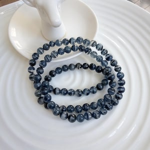 Op de afbeelding: Drie donkerblauwe kralenarmbanden met een gemarmerd effect liggen op een witte, geribbelde plaat. De kralen zijn klein en rond, met een mix van donkere en lichtblauwe tinten. De armbanden zijn in een cirkelvormige opstelling.