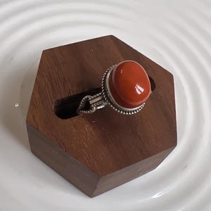 May include: A silver ring featuring a large, round, red gemstone set in a silver bezel. The ring is displayed on a wooden ring holder.