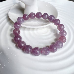 May include: A bracelet made of round, polished, purple gemstones. The beads are strung together to form a circular shape. The bracelet is resting on a white, decorative plate.